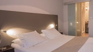 Una cama bien hecha con sábanas blancas y un plaid estampado, una lámpara de noche, y vista a un baño con ducha y toallero.