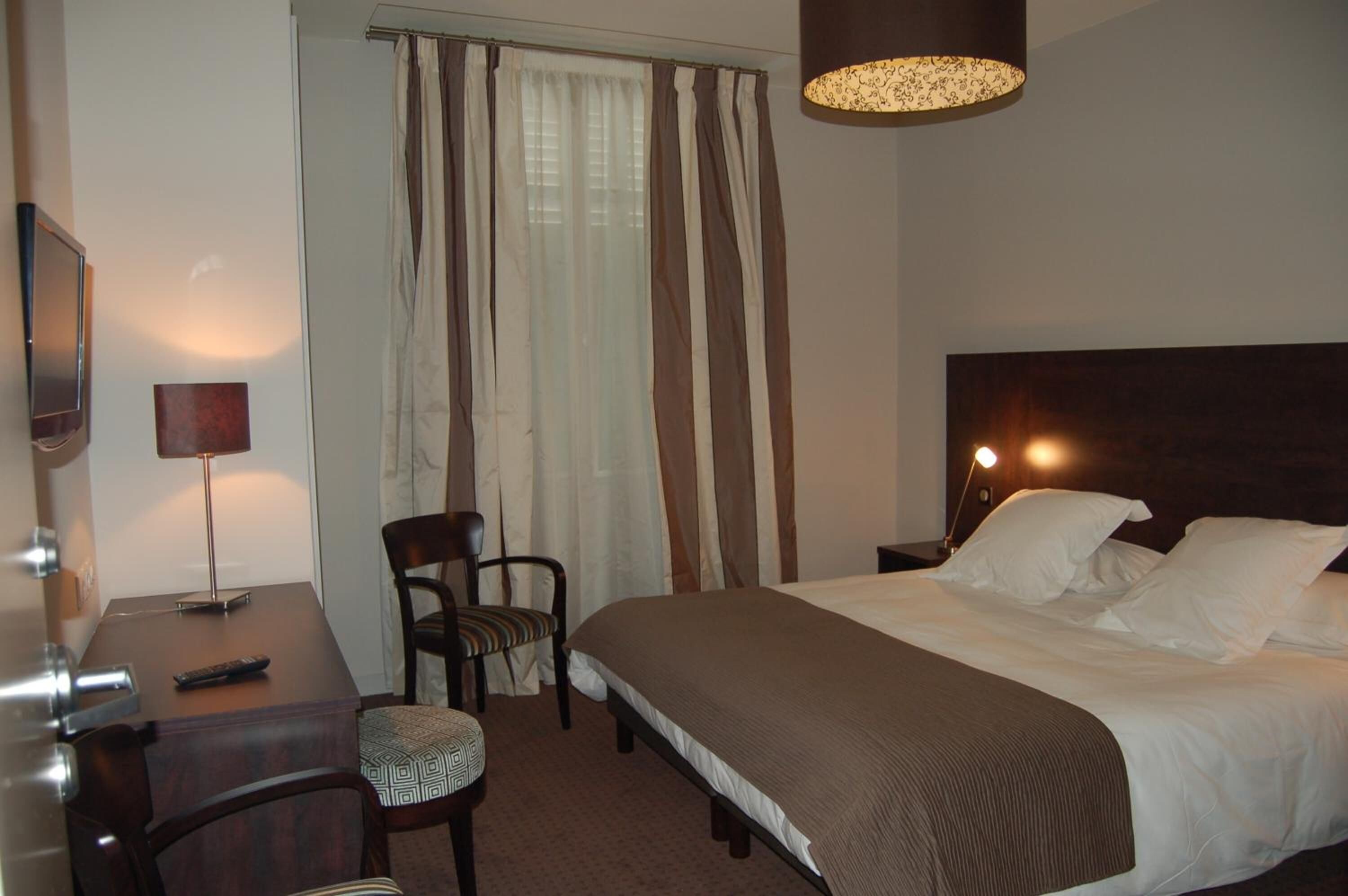 A hotel room with a bed, a desk with a lamp, a chair, and a TV.