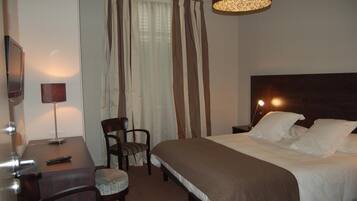 A hotel room with a bed, a desk with a lamp, a chair, and a TV.
