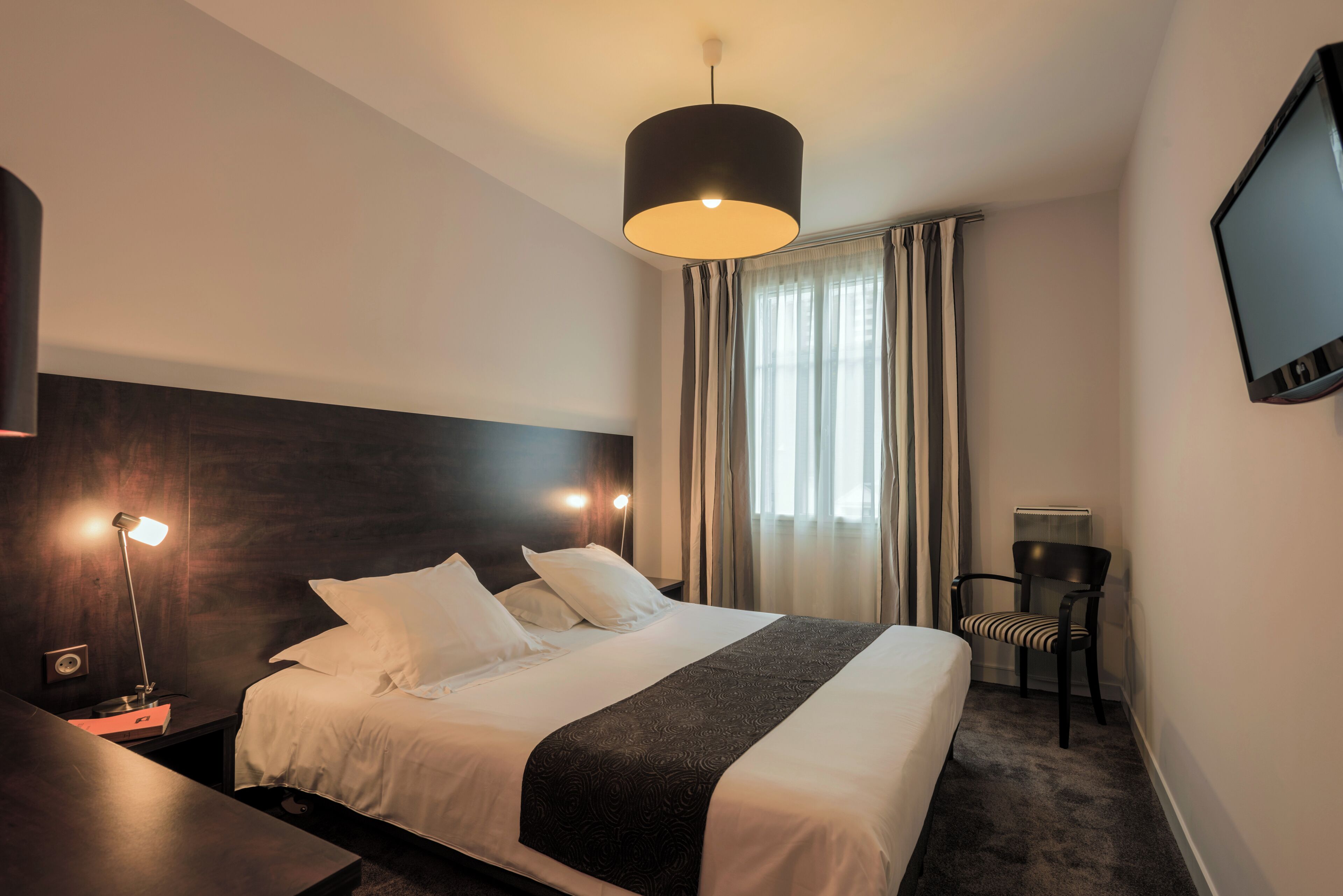 Ein Hotelzimmer mit einem Bett, Nachttischlampen, einem Fernseher, einem Sessel und einem Fenster mit Vorhängen.