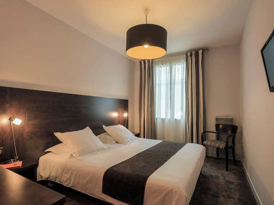 Ein Hotelzimmer mit einem Bett, Nachttischlampen, einem Fernseher, einem Sessel und einem Fenster mit Vorhängen.