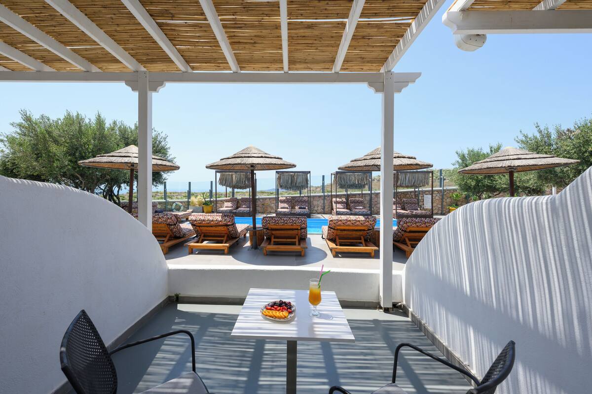 A covered outdoor seating area with wooden lounge chairs and thatched umbrellas, overlooking a pool and a scenic view.