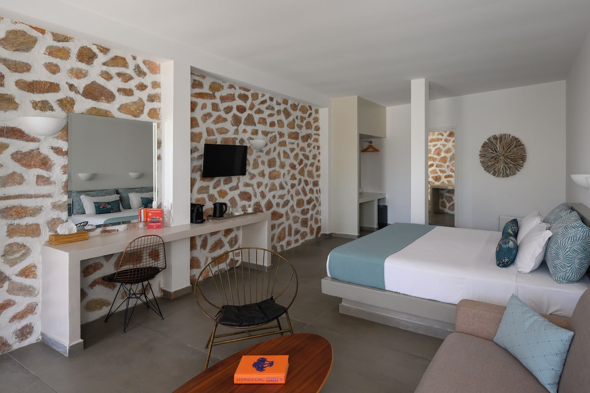 A modern hotel room with a stone-patterned wall, a bed with patterned pillows, a desk with a flat-screen TV, and a seating area with a coffee table and chairs.