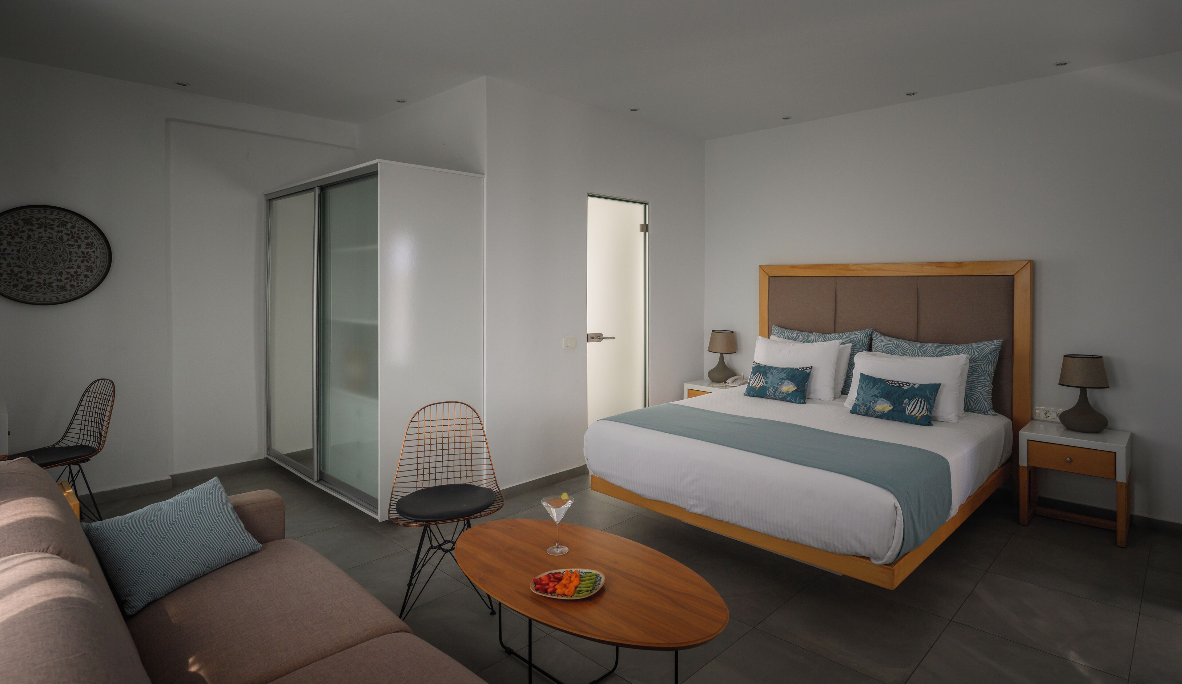 A modern hotel room with a large bed, a sofa, a dining table, and a sliding door leading to a balcony.