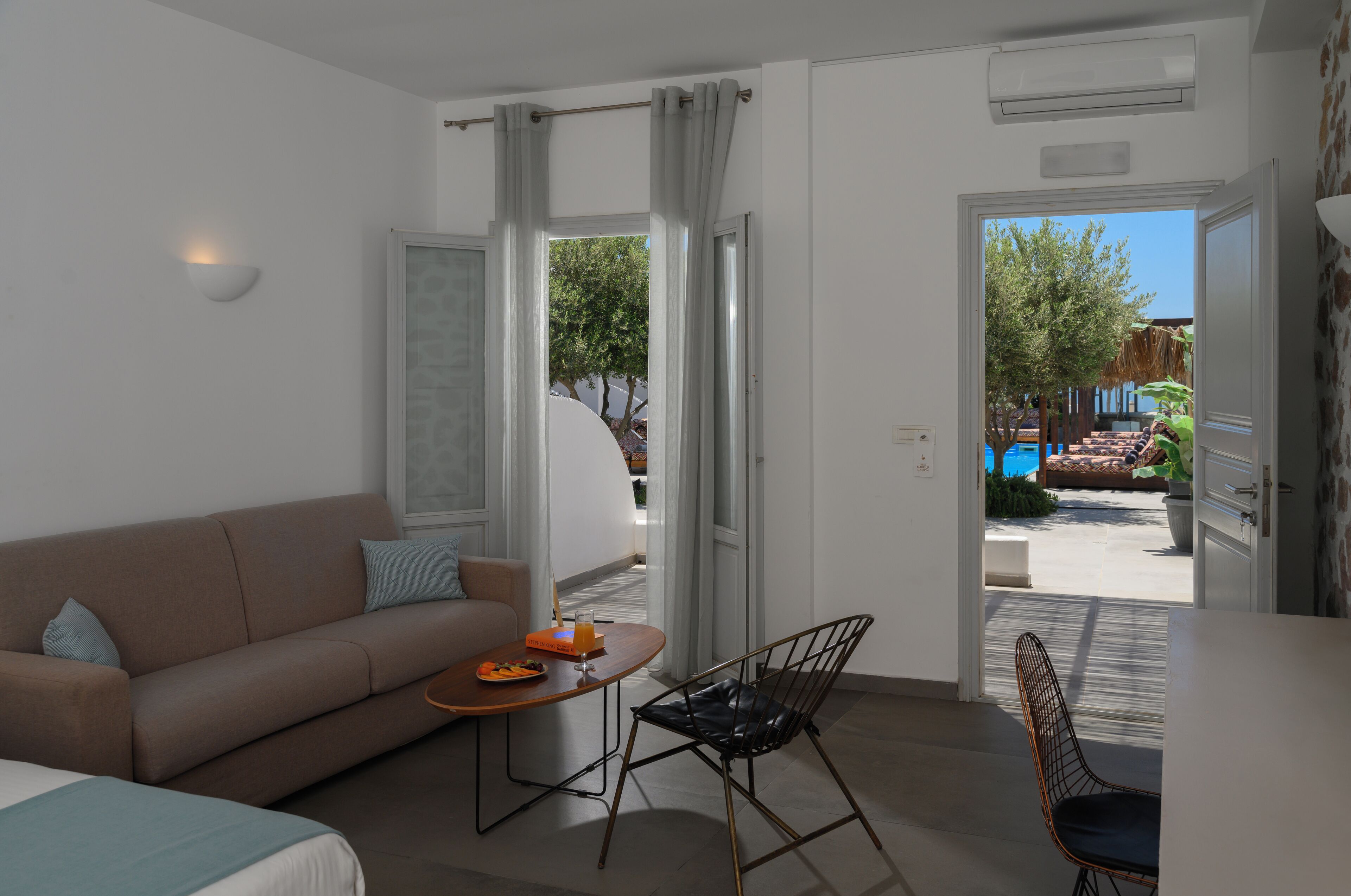 A modern hotel room with a sofa, a dining table with a fruit bowl, and a view of an outdoor pool area.