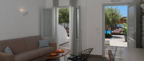 A modern hotel room with a sofa, a dining table with a fruit bowl, and a view of an outdoor pool area.