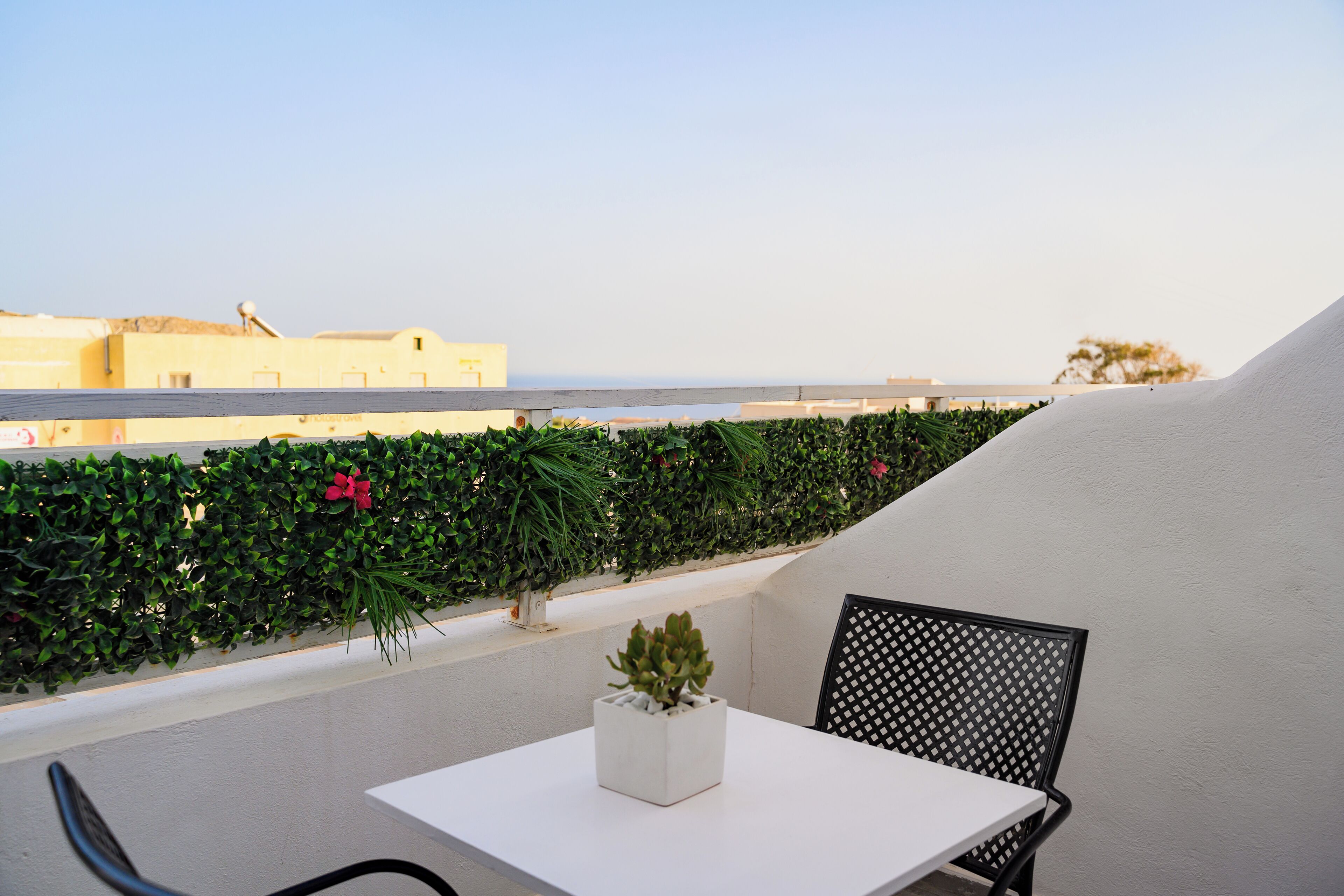 A balcony with a white table and a black chair, a potted plant, and a view of the sea and buildings in the background.
