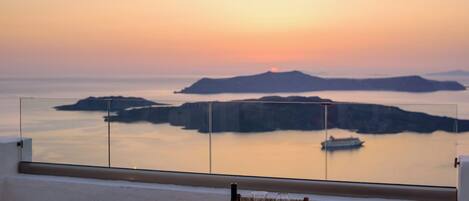 A round table with chairs on a balcony overlooking a sunset view of islands and a boat.