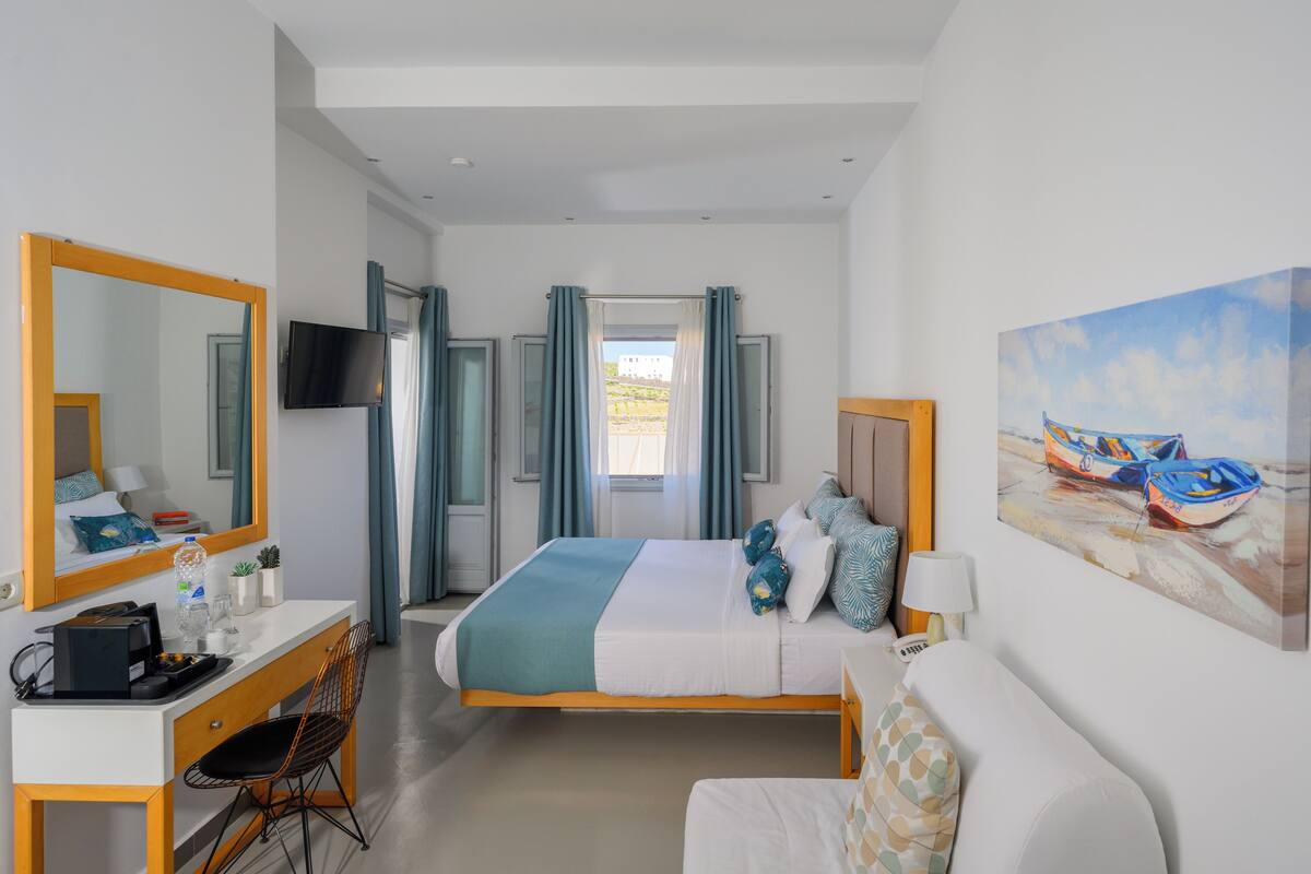 A hotel room with a bed, a desk, a chair, a TV, and a painting of boats on the wall.