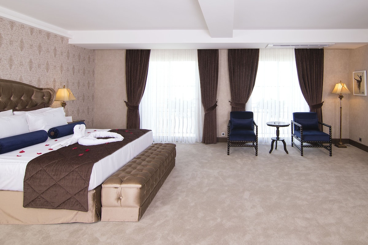 Una habitación de hotel con una cama grande, un banco, dos sillones, una mesita y un televisor.