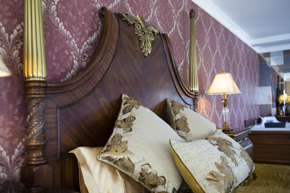 Una cama con cabecera decorada y almohadas estampadas, una mesita de noche con lámpara y un papel tapiz con diseño.
