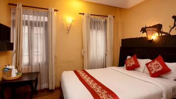 A hotel room with a bed, curtains, a wall-mounted light, a headboard with decorative pillows, and a small table with a basket and bottle.