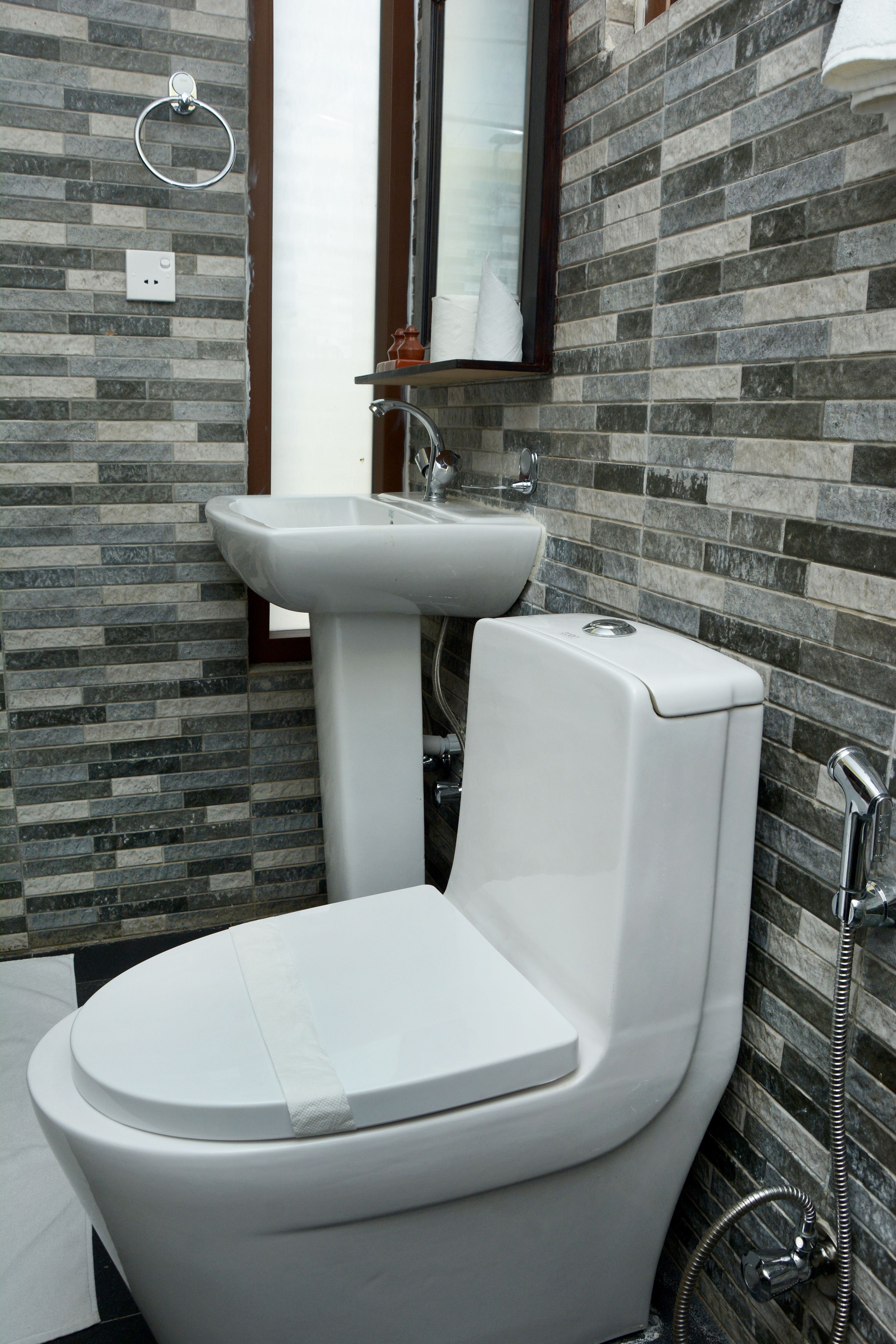 A bathroom with a white toilet, a pedestal sink, and a shower. The walls are tiled with a mix of gray and brown tiles.