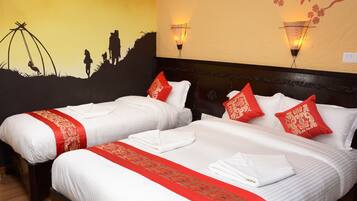A hotel room with two beds, each with white linens and red decorative pillows. There are wall-mounted lights and a mural depicting a silhouette of people on a hill.