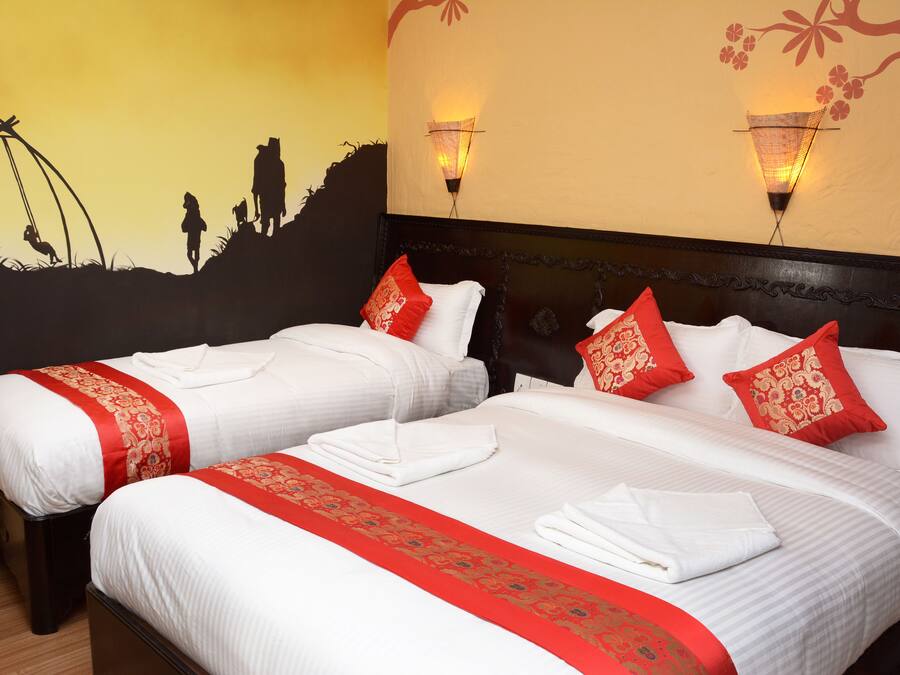 A hotel room with two beds, each with white linens and red decorative pillows. There are wall-mounted lights and a mural depicting a silhouette of people on a hill.