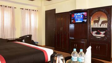 A hotel room with a bed, a television, a cabinet with a deer decoration, and a table with a kettle and water bottles.