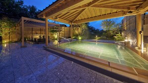 A covered outdoor swimming pool with a wooden roof, surrounded by stone walls and a tiled floor.