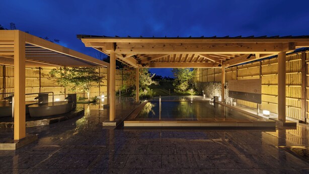 A covered outdoor pool area with a wooden pergola, stone flooring, and a seating area with a hot tub.