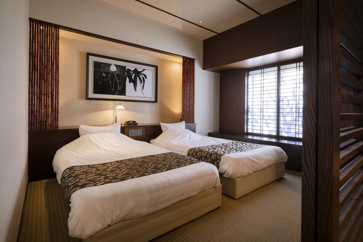 A hotel room with two beds, a framed picture on the wall, and a window with blinds.