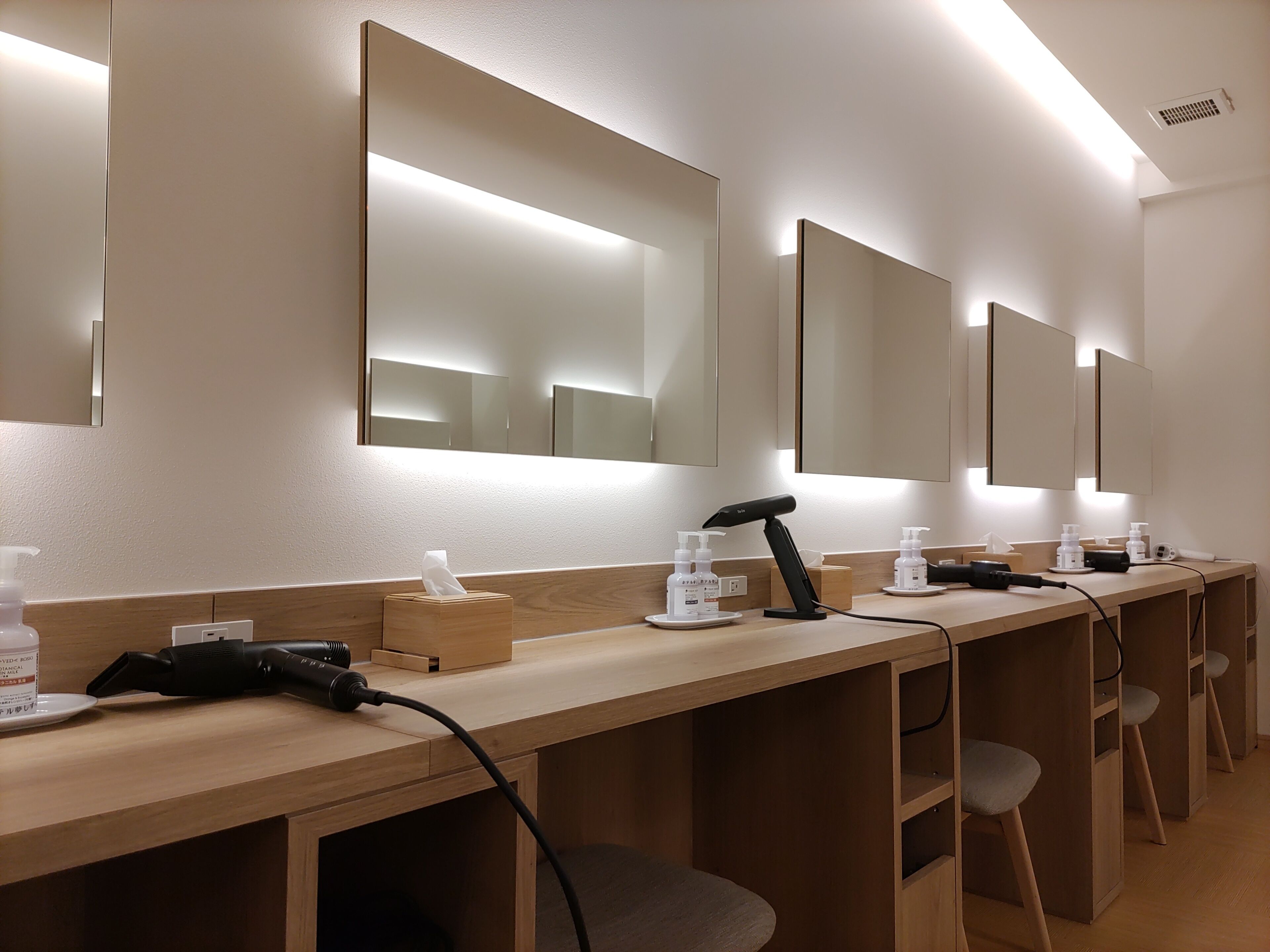 A row of hair dryers and personal care items on a wooden counter in a well-lit room with mirrors on the wall.