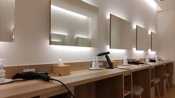 A row of hair dryers and personal care items on a wooden counter in a well-lit room with mirrors on the wall.