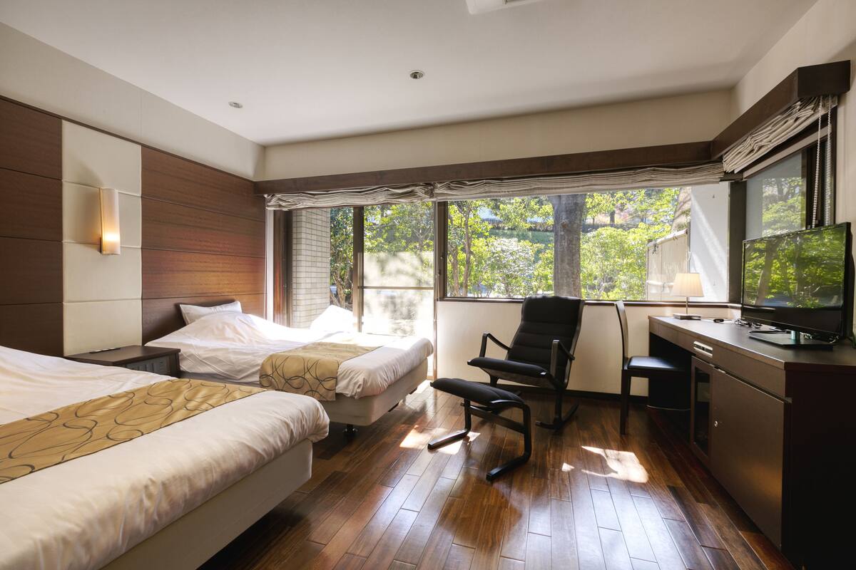 A hotel room with two beds, a desk with a television, and large windows offering a view of greenery.