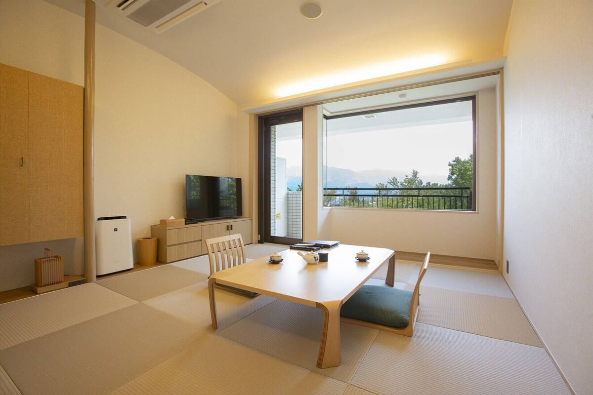 A room with tatami flooring, a low table with cushions, a television on a cabinet, and a large window with a view of mountains.
