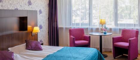 A hotel room with a bed, two chairs, a small table, and a lamp.