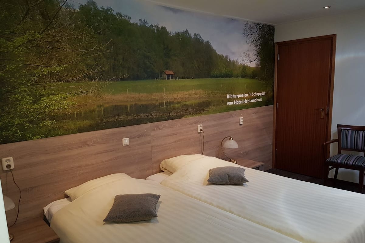 Une chambre d’hôtel avec deux lits, une tête de lit en bois, une fresque représentant un paysage et une chaise.