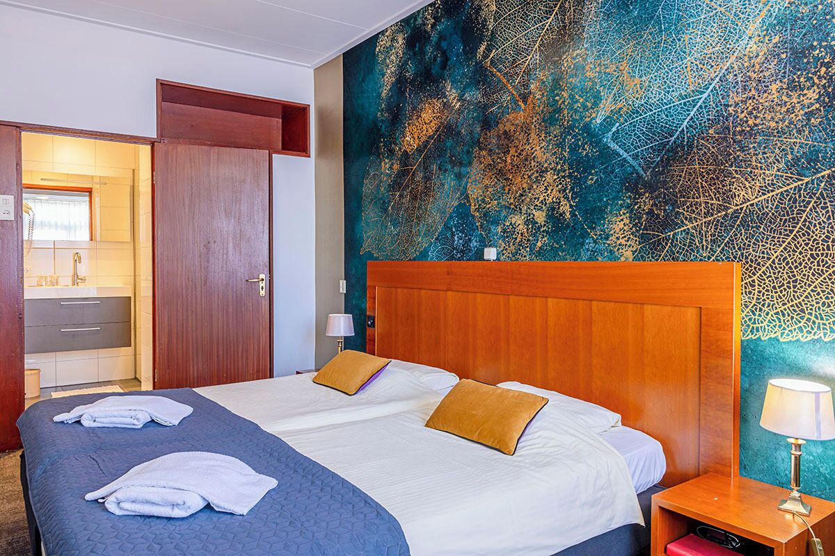 Hotellihuone, jossa on suuri sänky, kaksi tyynyä, yöpöytä lampun kanssa ja seinä, jossa on lehtikuvio.