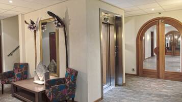 A lobby with a reception desk, two floral armchairs, a large mirror, and an elevator.