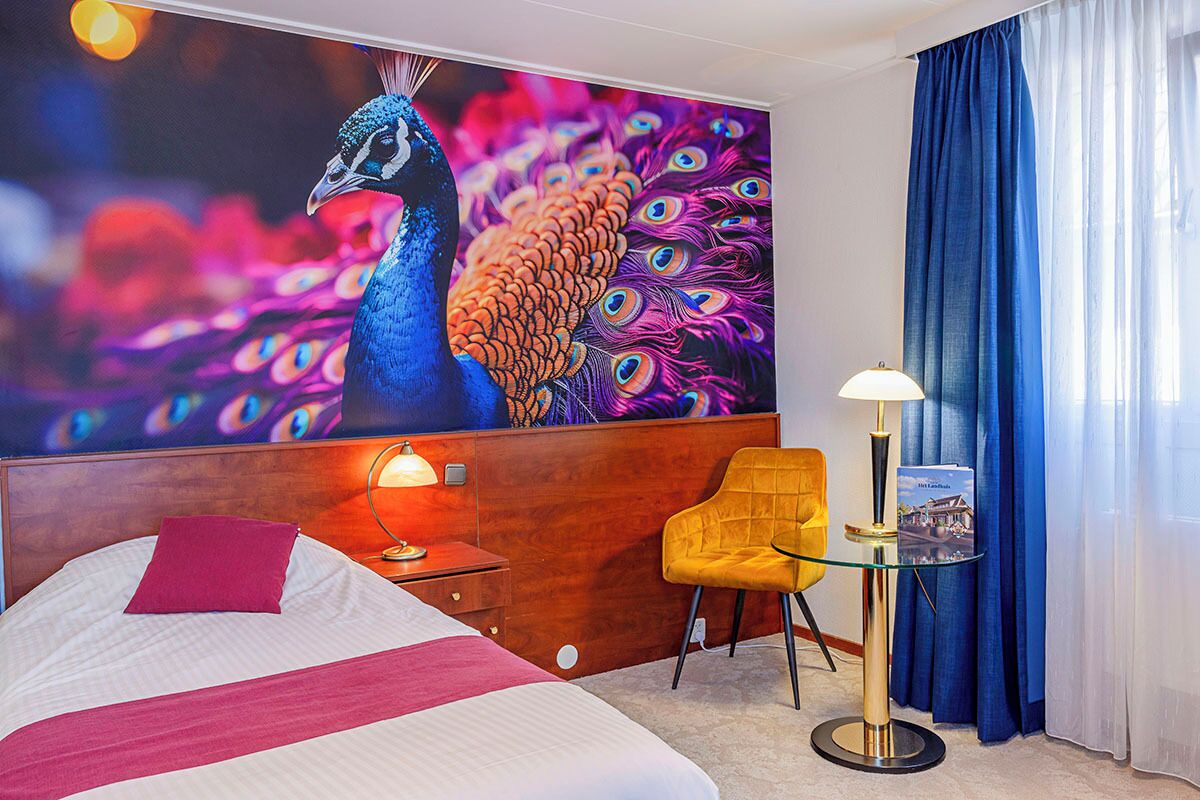 A hotel room with a large peacock mural, a bed with a red pillow, a yellow armchair, a small table with a lamp, and a view of the outside through sheer curtains.