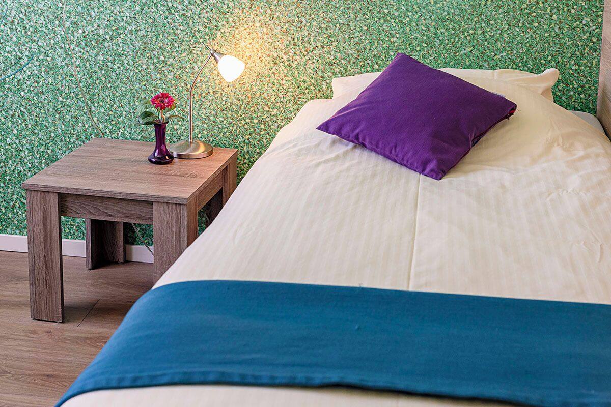 Un lit bien fait, recouvert d’une couvre-lit bleue et blanche, avec un oreiller violet et une petite table de chevet en bois sur laquelle se trouvent une lampe et un vase avec une fleur.