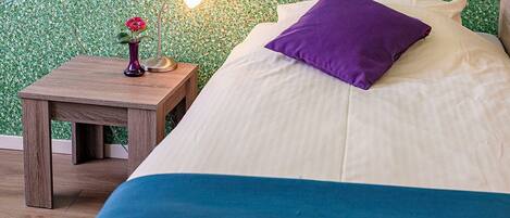 Una cama bien hecha con colcha azul y blanca, un cojín morado y una pequeña mesita de noche con una lámpara y un florero con una flor.