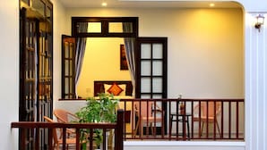 Uma sala de estar acolhedora com sofá, cadeiras e uma mesa de jantar. Há uma varanda com guarda-corpo e plantas em vasos.