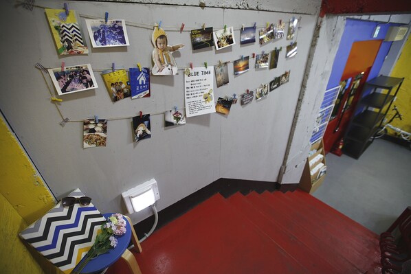 A room with a red carpeted staircase, a wall adorned with various photographs and a 'BLUE TIME' sign, and a table with a patterned tablecloth and a bouquet of flowers. - Bluefish Guesthouse - Hostel (Busan)