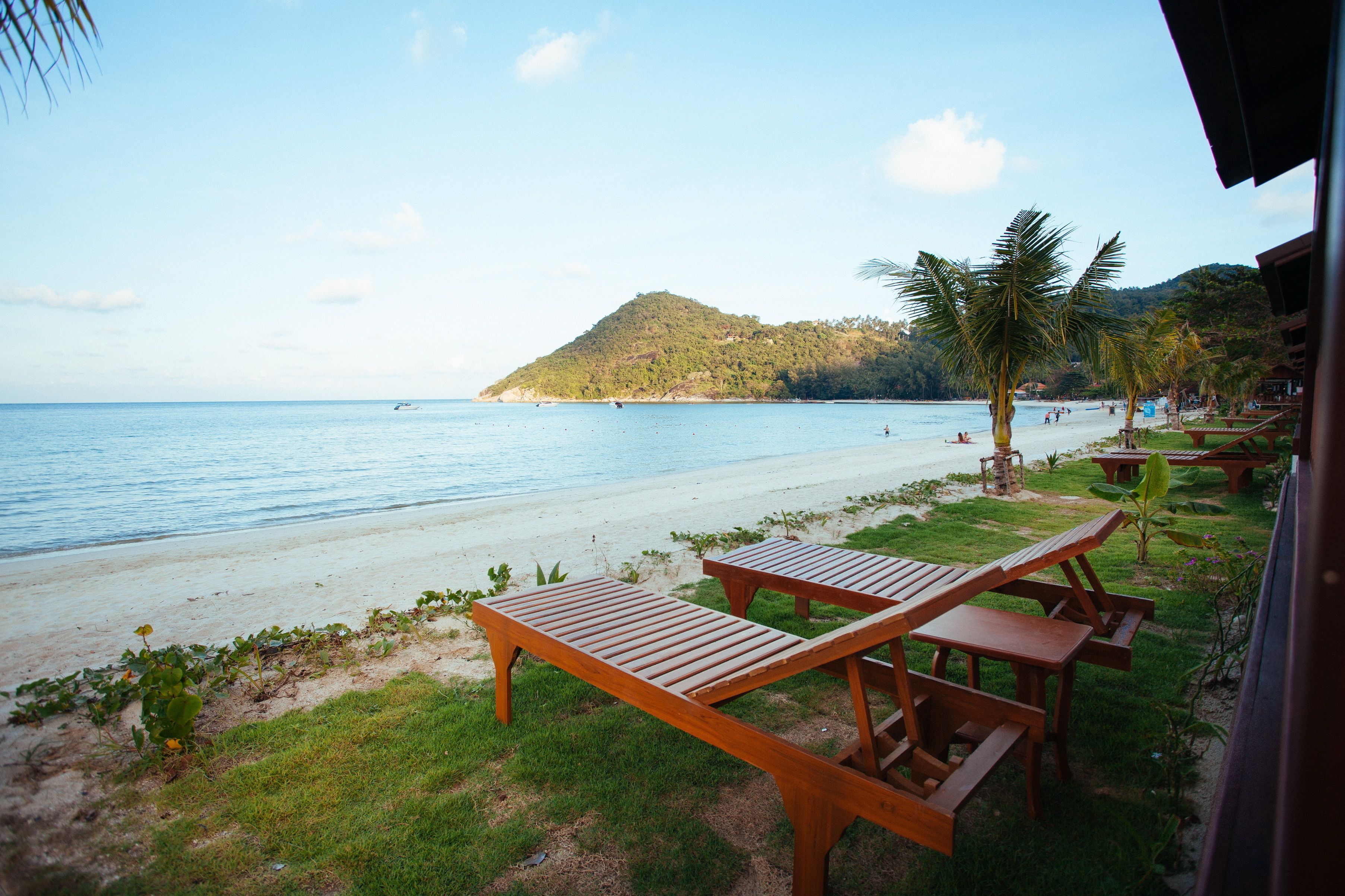 A beach with wooden benches and tables, a clear view of the sea, and a hill in the background.