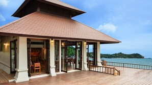 Un espace extérieur abrité doté d’une terrasse en bois, de portes vitrées donnant sur un espace intérieur aménagé avec des sièges, et offrant une vue sur la mer.