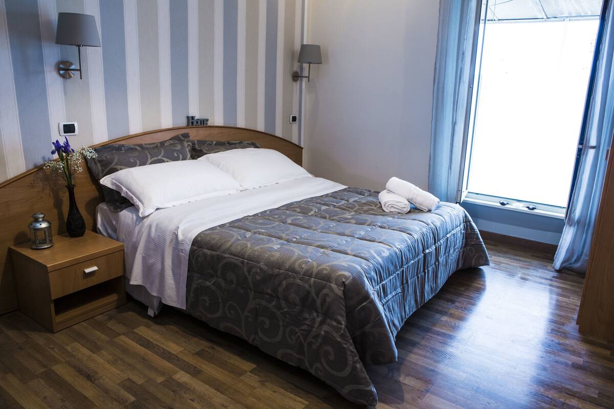 A neatly made bed with a patterned bedspread, a bedside table with a lamp, and a nightstand with a vase and a lantern.