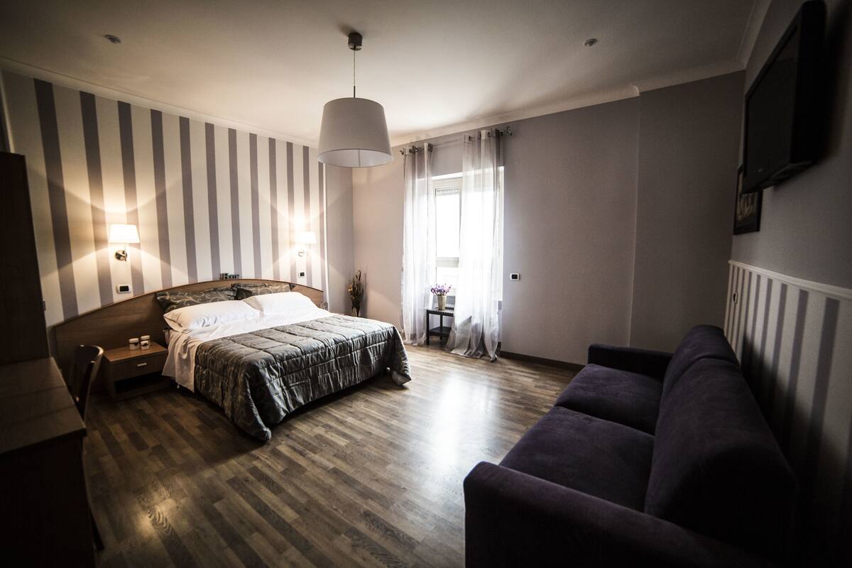 A hotel room with a bed, a sofa, a desk, a lamp, and a window with curtains.