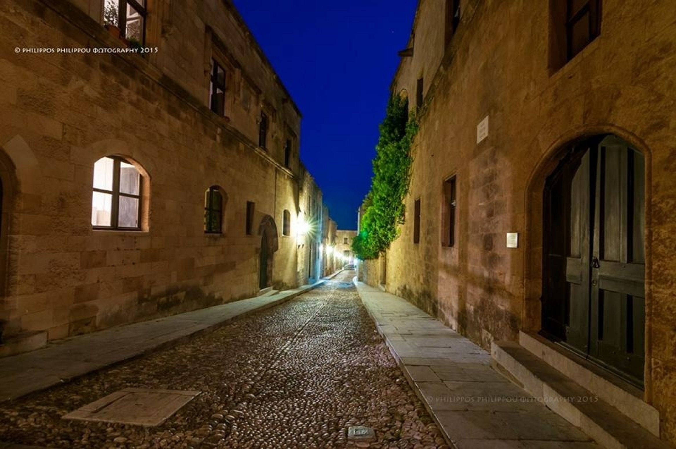 Une ruelle étroite pavée, bordée de bâtiments anciens en pierre dotés d’arcs au-dessus des fenêtres et des portes, qui mène à un ciel sombre.