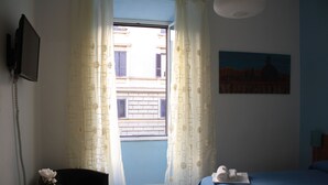 A hotel room with a bed, a chair, a small table with a plant, a painting on the wall, and a window with curtains. - Scipione B&B (Rome)
