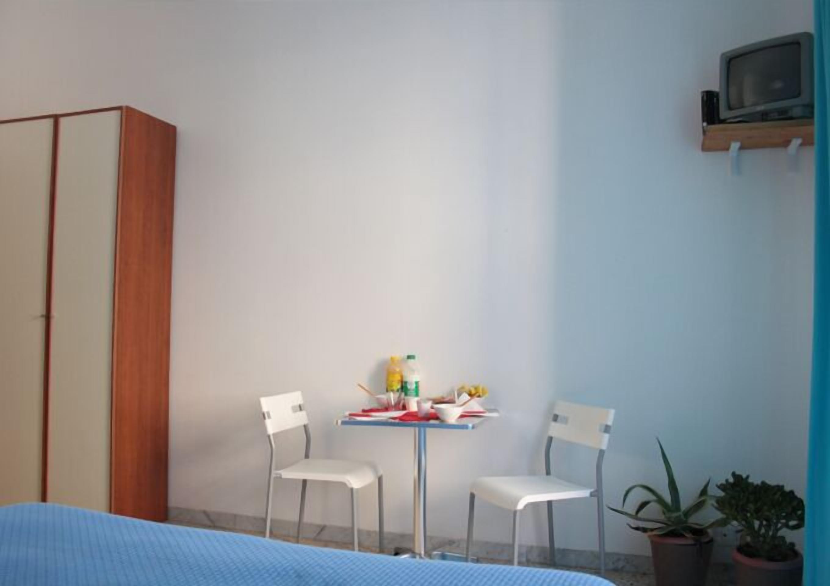 A room with a bed, two white chairs, a small table with food items, a TV on a shelf, and two potted plants.