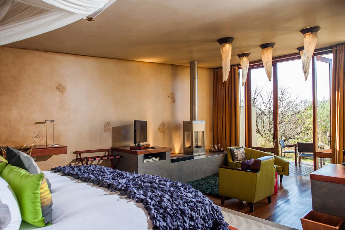 Una habitación de hotel moderna con una cama grande, una zona de estar con sofá y sillones, una chimenea y ventanas amplias con vistas al exterior.