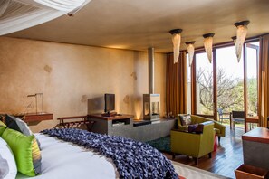 A modern hotel room with a large bed, a seating area with a sofa and chairs, a fireplace, and large windows offering a view of the outdoors. - Forum Homini Boutique Hotel (Krugersdorp)