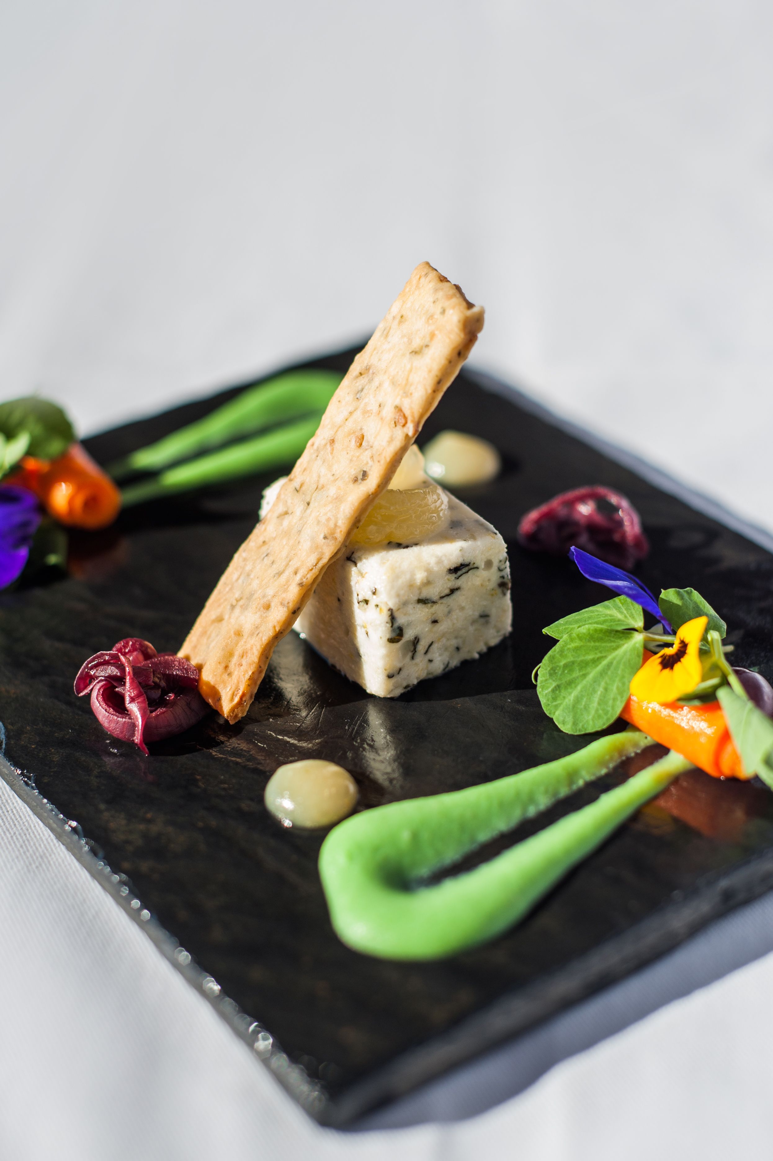 A square plate with a piece of blue cheese, a cracker, and various garnishes including a green leaf, a purple flower, and a yellow flower.