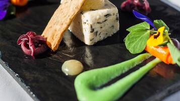 A square plate with a piece of blue cheese, a cracker, and various garnishes including a green leaf, a purple flower, and a yellow flower.