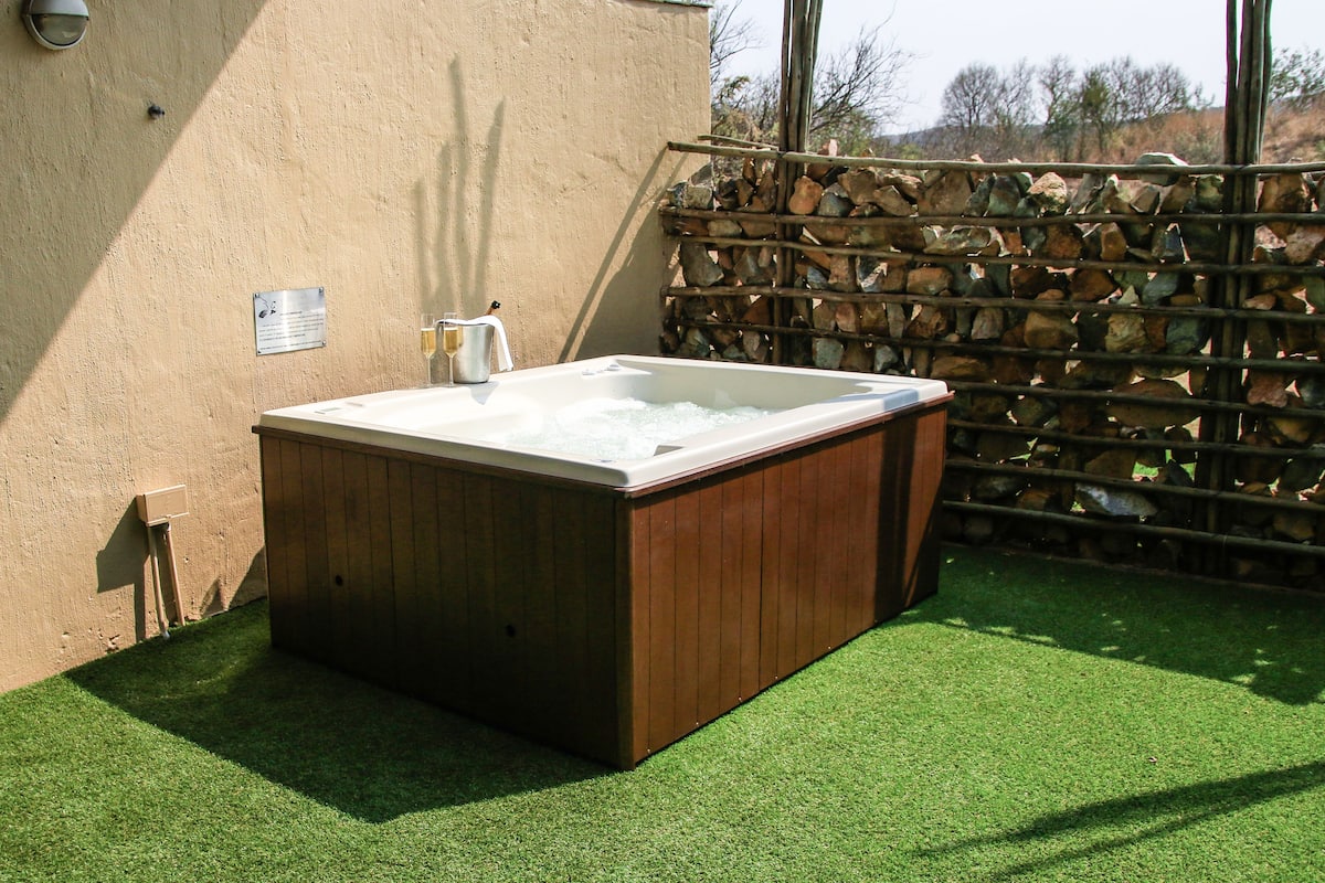 Un jacuzzi con revestimiento de madera, ubicado frente a una pared con cerramiento de piedra y césped artificial.