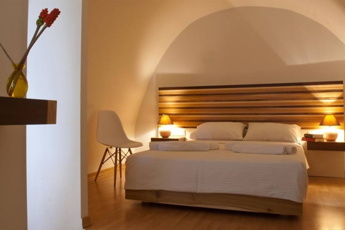 Een slaapkamer met een houten hoofdeinde, een nachtkastje met een lamp, een stoel en een plank met wijnflessen en bloemen.