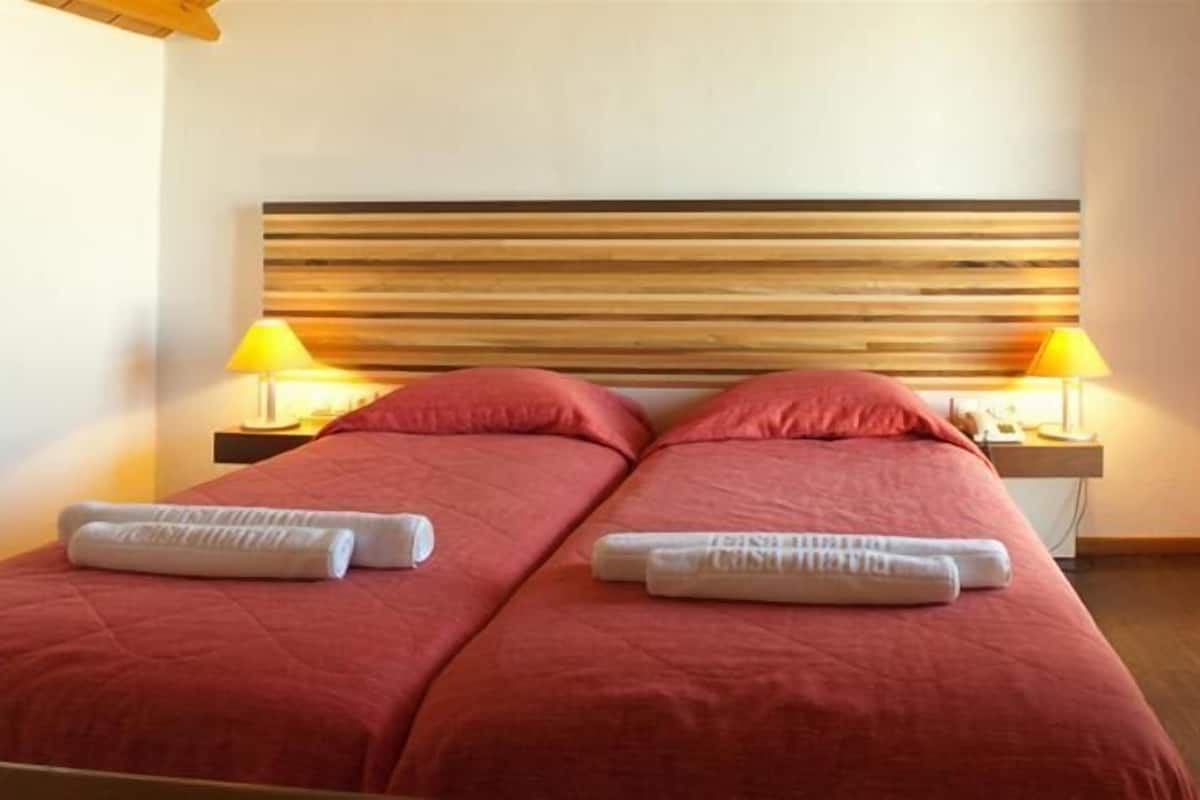 Een tweepersoonskamer met twee bedden, elk met een rode sprei en witte handdoeken. Er staan twee nachtkastjes met lampen op, en links is een stoel te zien.